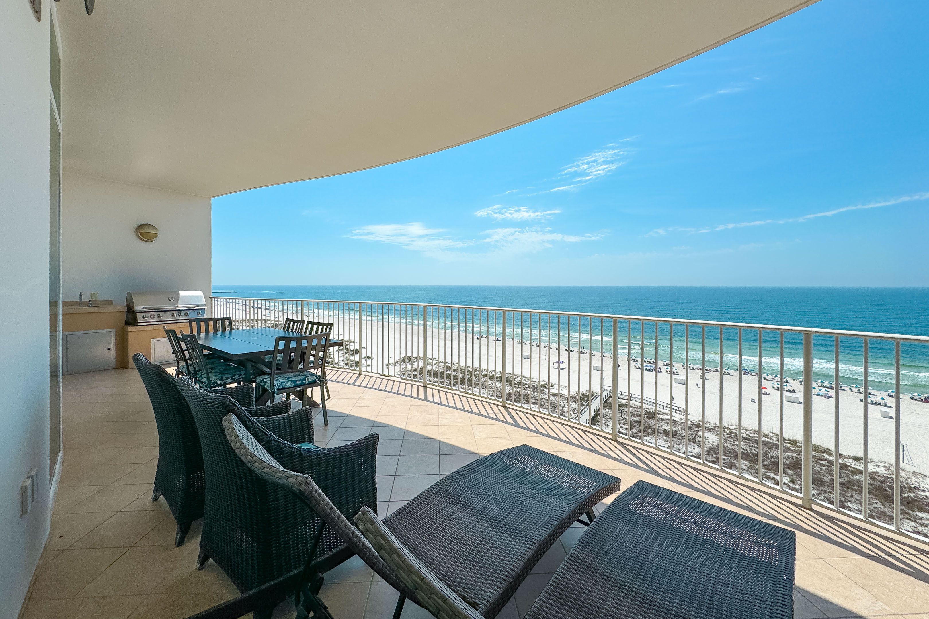 A spacious beachfront balcony with lounge chairs, a dining table, and a built-in grill station, overlooking white sand beaches and the turquoise waters of the Gulf under a clear blue sky.