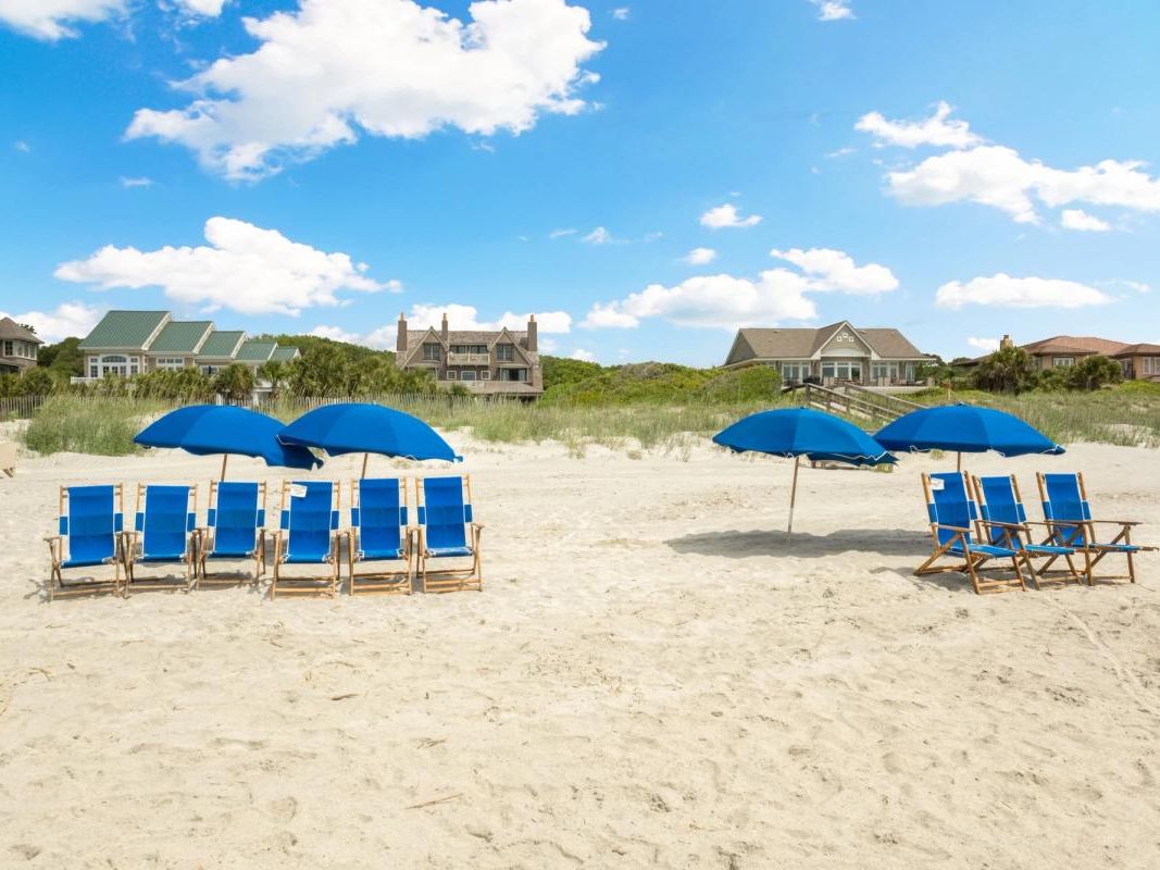 Rows of bright blue beach chairs and umbrellas lined up on a wide, sandy beach with beach homes in the background. A perfect spot for families to relax by the ocean in Kiawah Island.