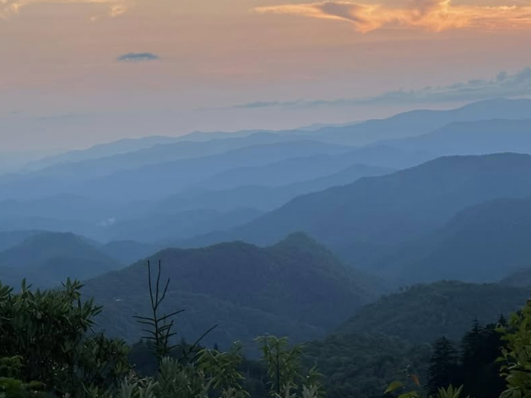 A scenic view of rolling mountain ridges at sunset, with layers of blue-toned peaks fading into the distance. The sun is partially hidden behind clouds, casting a warm orange glow across the sky. Green foliage in the foreground adds depth to the tranquil and picturesque landscape