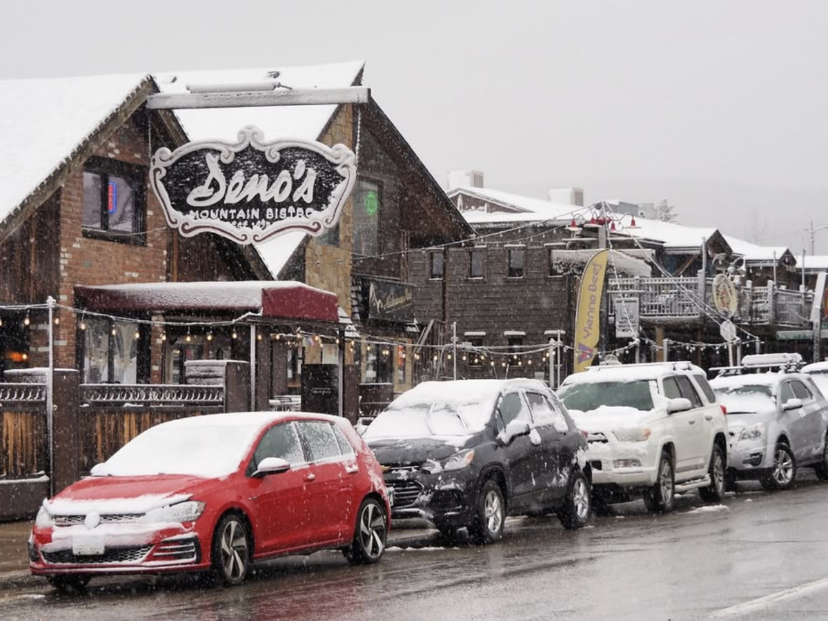 Snow-covered cars line the street in front of Deno's Mountain Bistro and other rustic buildings during a snowy day in a charming mountain town.