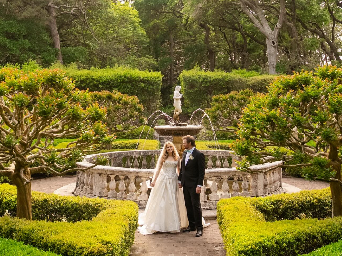 A bride and groom stand hand in hand before a stone fountain in a lush formal garden surrounded by manicured hedges and sculpted trees.