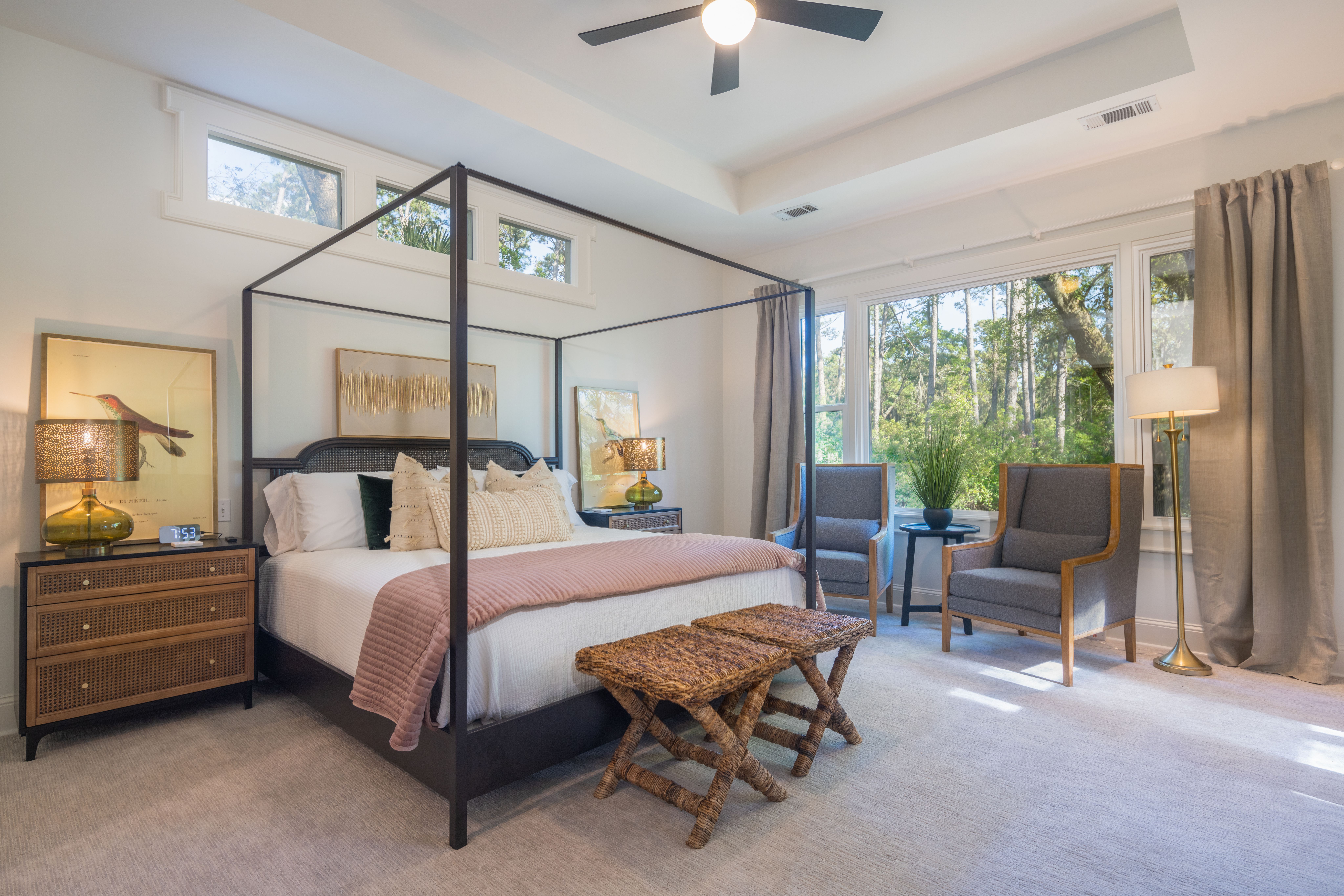 A serene bedroom features a four-poster bed with soft bedding and decorative pillows, flanked by nightstands with glass lamps, and includes a cozy sitting area with two armchairs by large windows overlooking a wooded view.
