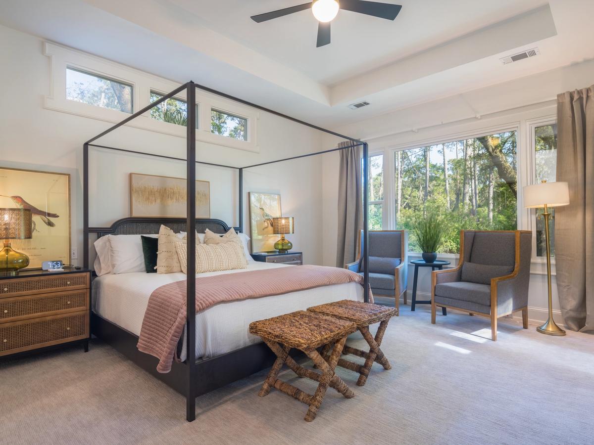 A serene bedroom features a four-poster bed with soft bedding and decorative pillows, flanked by nightstands with glass lamps, and includes a cozy sitting area with two armchairs by large windows overlooking a wooded view.
