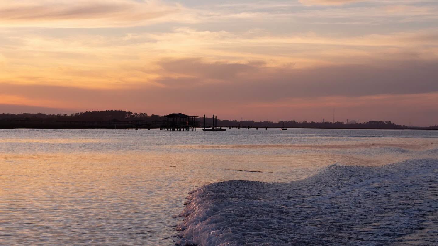  A calm tidal river glows in soft pastel shades of orange, pink, and lavender at sunset, with a distant dock and tree-lined shore silhouetted on the horizon. The wake of a passing boat ripples across the still water in the foreground
