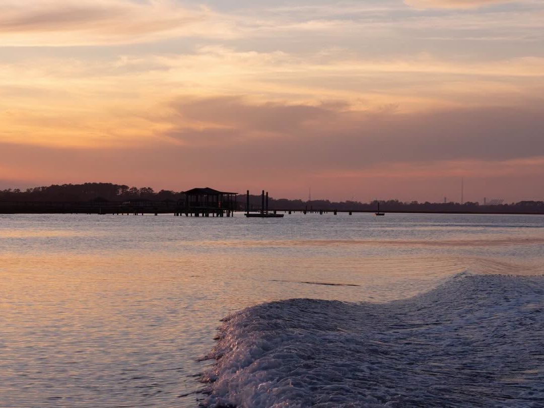 A calm tidal river glows in soft pastel shades of orange, pink, and lavender at sunset, with a distant dock and tree-lined shore silhouetted on the horizon. The wake of a passing boat ripples across the still water in the foreground