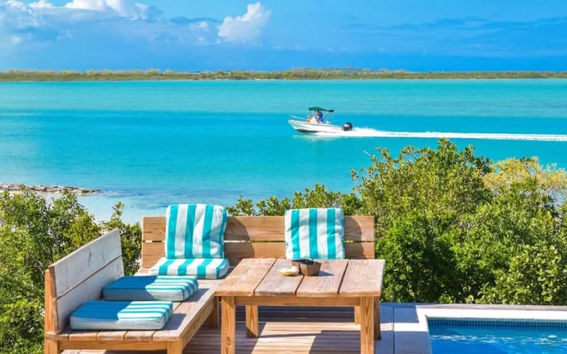 A cozy outdoor seating area with striped turquoise cushions overlooks a pool and bright blue ocean, where a boat speeds by in the distance.