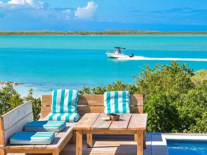 A cozy outdoor seating area with striped turquoise cushions overlooks a pool and bright blue ocean, where a boat speeds by in the distance.