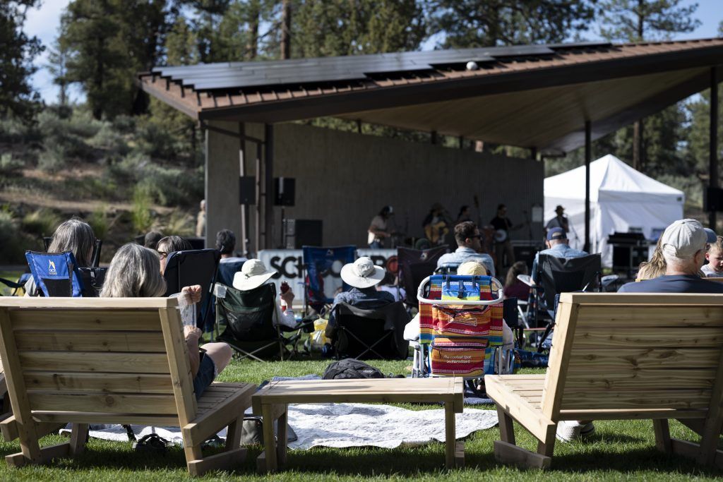 People relax on lawn chairs and wooden benches while enjoying live music under a modern outdoor pavilion surrounded by tall pines. The casual setup, sunshine, and laid-back crowd capture the welcoming vibe of Bend’s Alpenglow Concert Series.