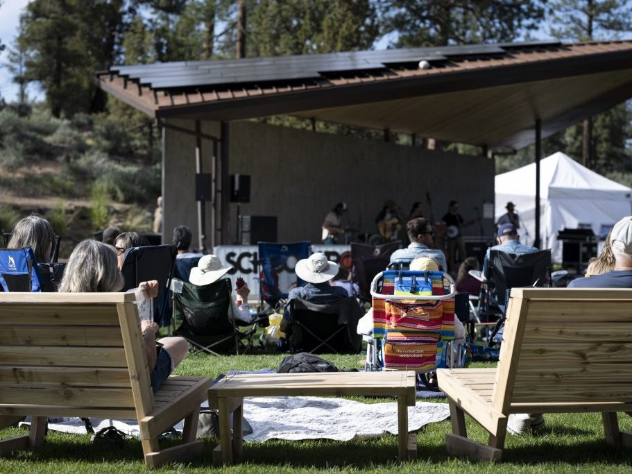 People relax on lawn chairs and wooden benches while enjoying live music under a modern outdoor pavilion surrounded by tall pines. The casual setup, sunshine, and laid-back crowd capture the welcoming vibe of Bend’s Alpenglow Concert Series.
