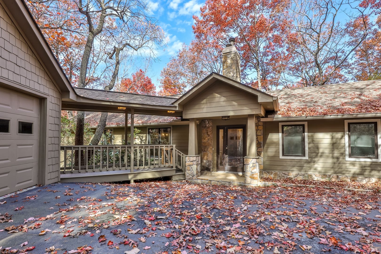 A charming single-level mountain cottage with stone accents, a covered entry and walkway, and neutral siding, surrounded by autumn foliage and fallen leaves in a wooded setting.