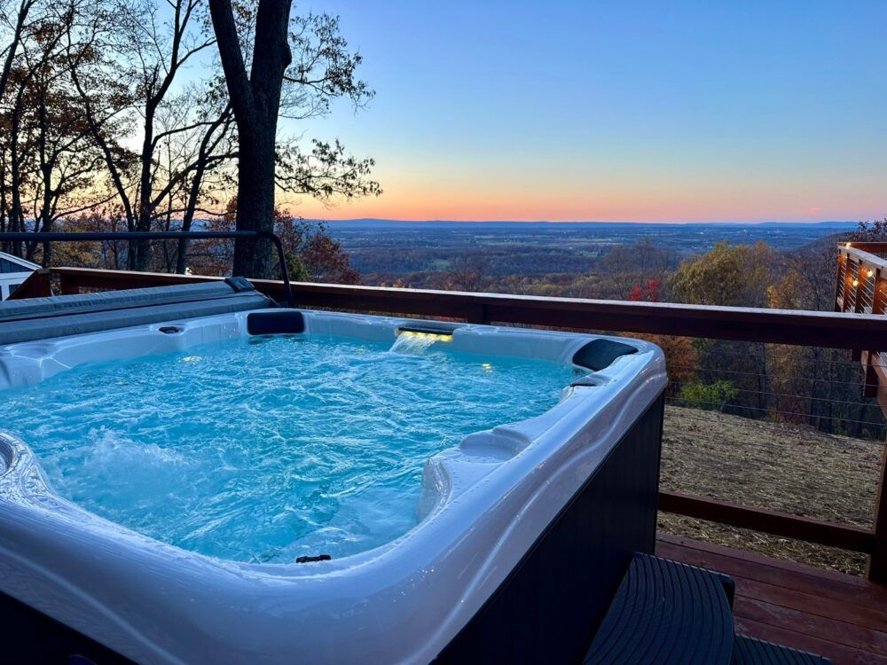 A steaming hot tub on a wooden deck overlooking a breathtaking panoramic mountain and valley view at sunset, with a glowing sky and autumn trees below.