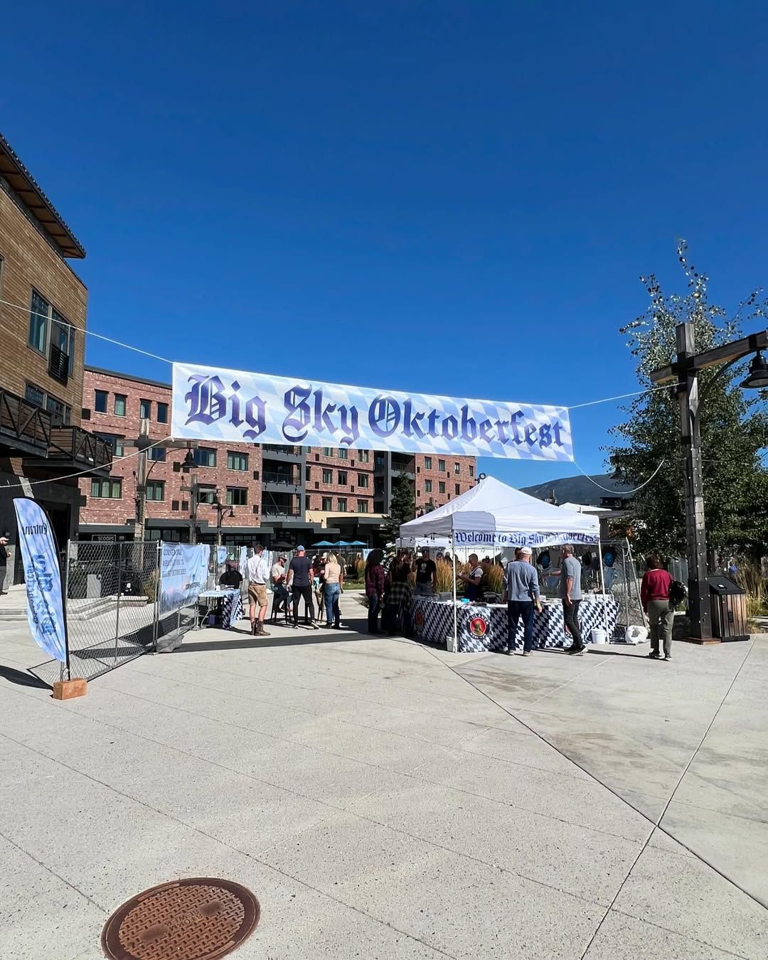 Visitors gather under a sunny blue sky for the Big Sky Oktoberfest, surrounded by mountain town buildings and festive tents. The cheerful event brings together food, drinks, and local fun in a lively outdoor plaza.