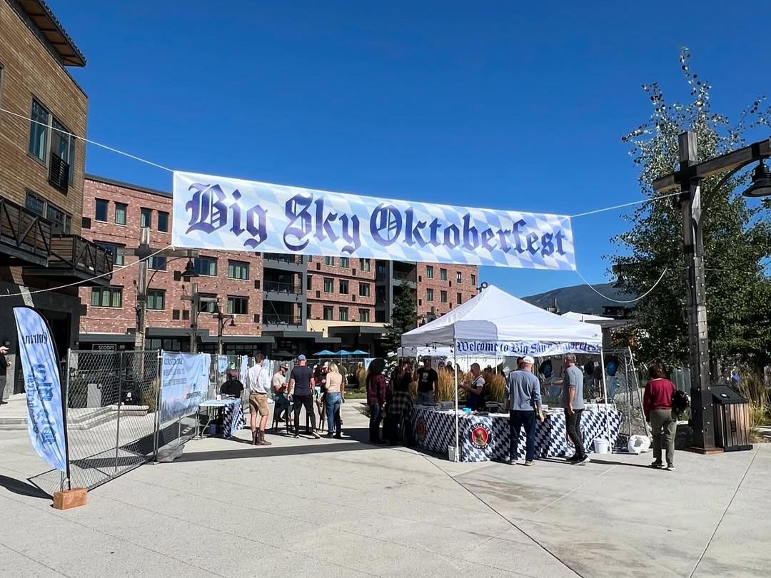 Visitors gather under a sunny blue sky for the Big Sky Oktoberfest, surrounded by mountain town buildings and festive tents. The cheerful event brings together food, drinks, and local fun in a lively outdoor plaza.