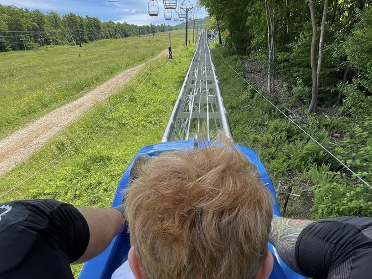 A child and adult ride an alpine coaster uphill through a lush green landscape with ski lift chairs hanging overhead.