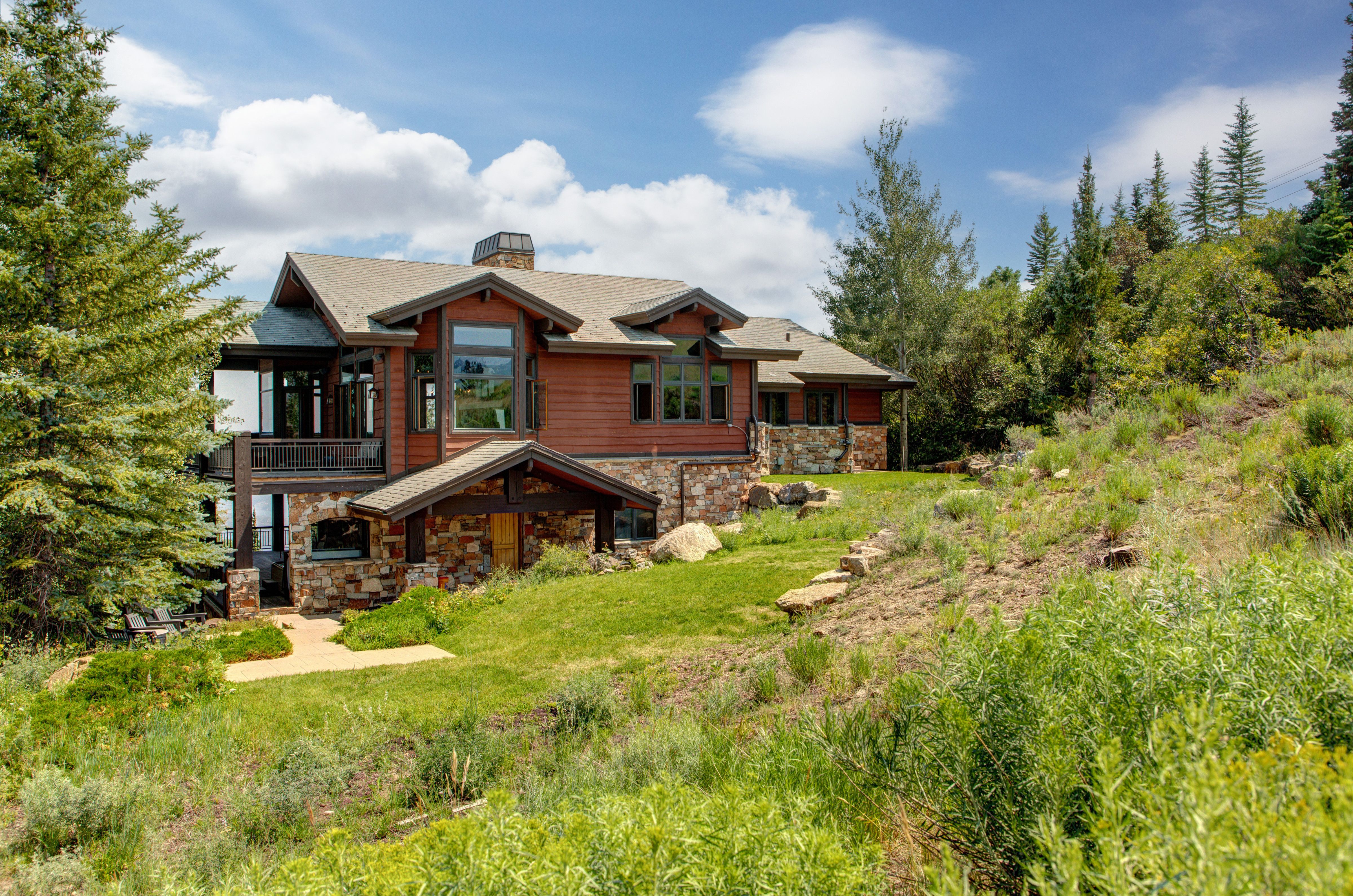 A rustic mountain home with wood siding, stone accents, and large windows, nestled in lush greenery with a backdrop of pine trees and a partly cloudy sky.