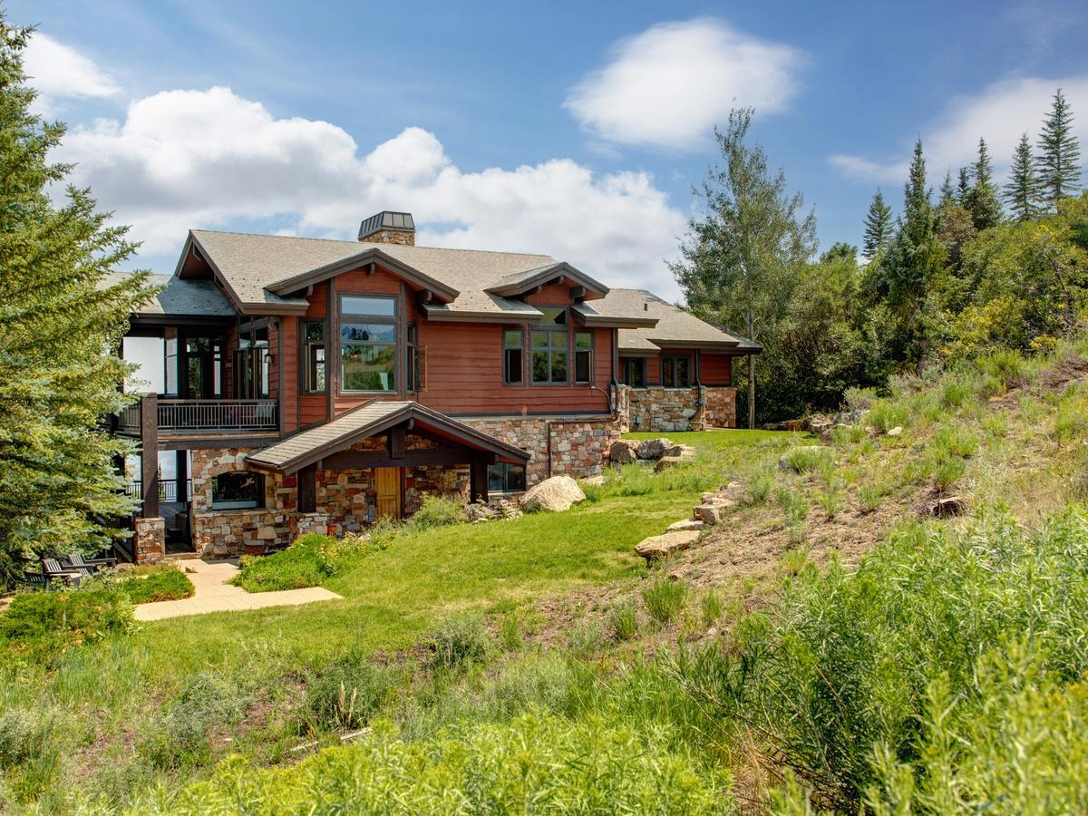 A rustic mountain home with wood siding, stone accents, and large windows, nestled in lush greenery with a backdrop of pine trees and a partly cloudy sky.