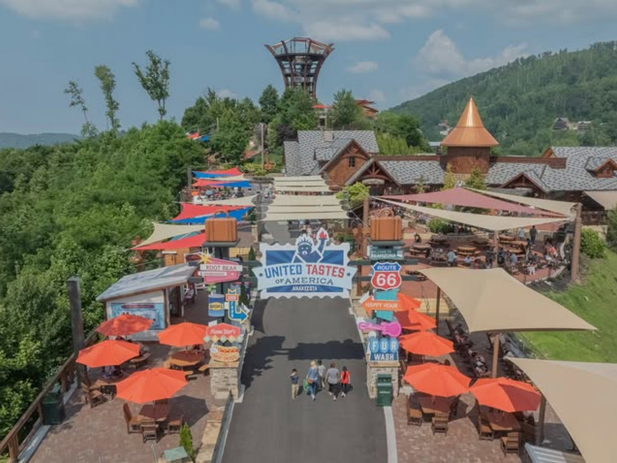 Visitors stroll through a lively outdoor plaza called “United Tastes of America,” lined with colorful umbrellas, food stands, and mountain views in the background.