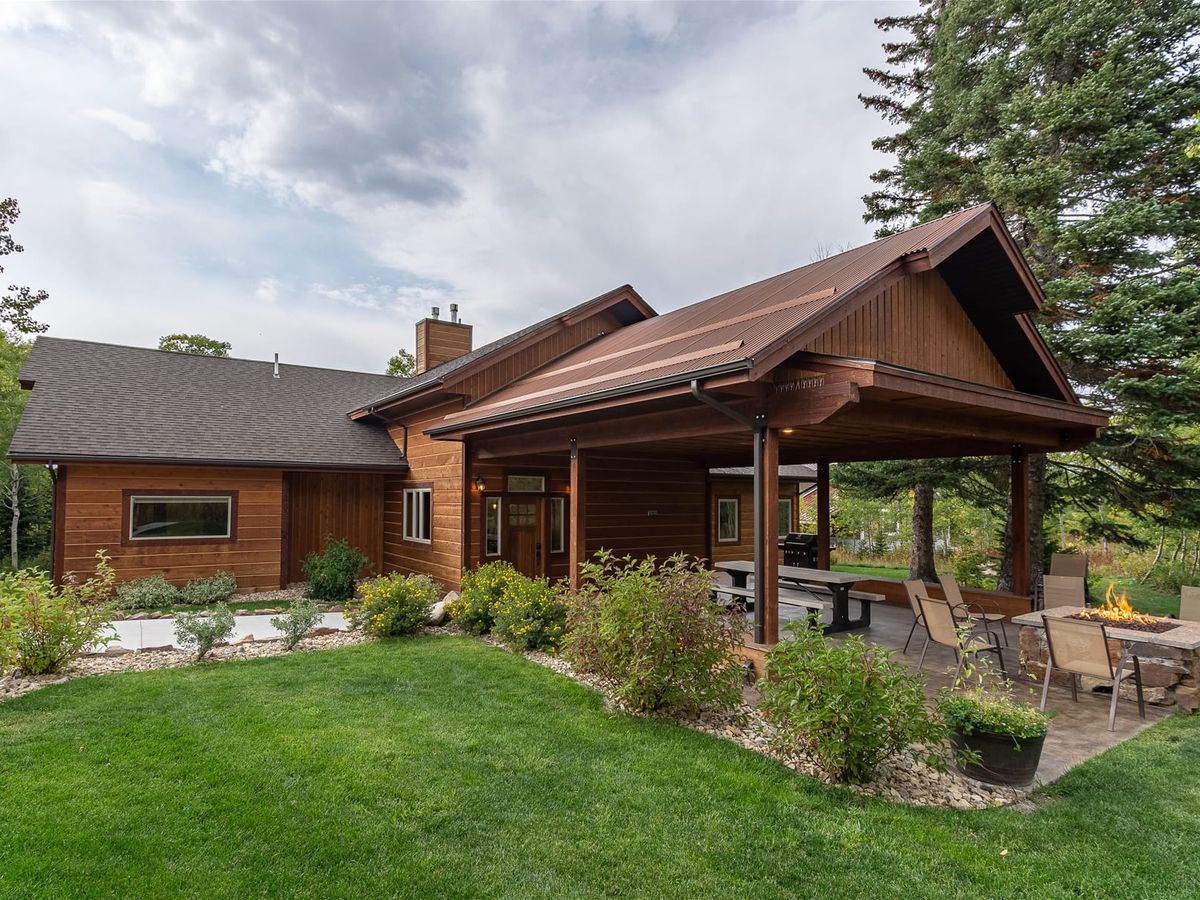 ChatGPT said: A cozy wooden cabin with a covered porch sits among lush greenery, featuring a patio area with outdoor dining, a fire pit, and surrounding gardens beneath tall pine trees.