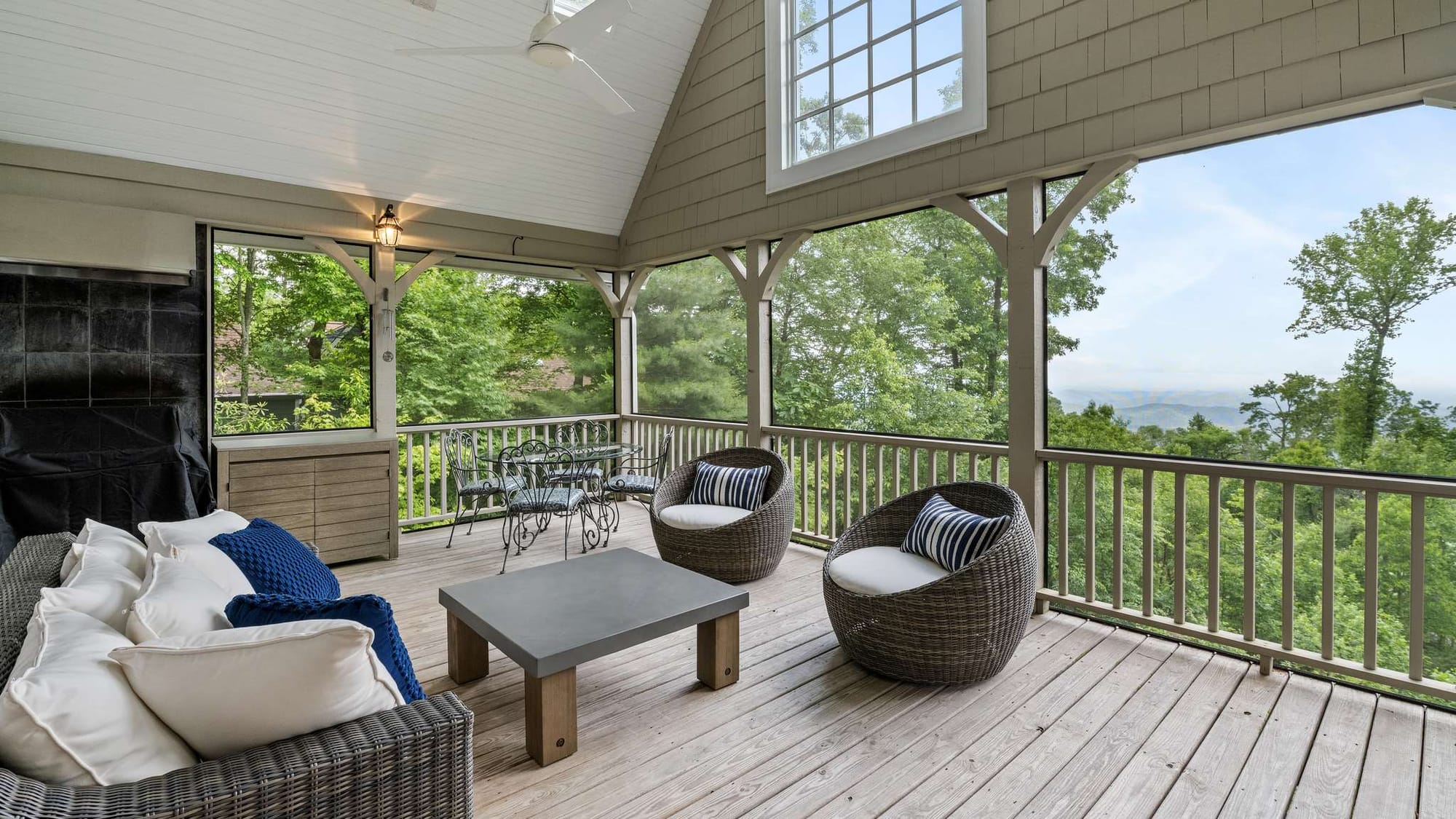A covered mountain porch features cozy seating and a dining table with wide views of the forest and distant hills. This outdoor living space is ideal for morning coffee or quiet evenings surrounded by nature.