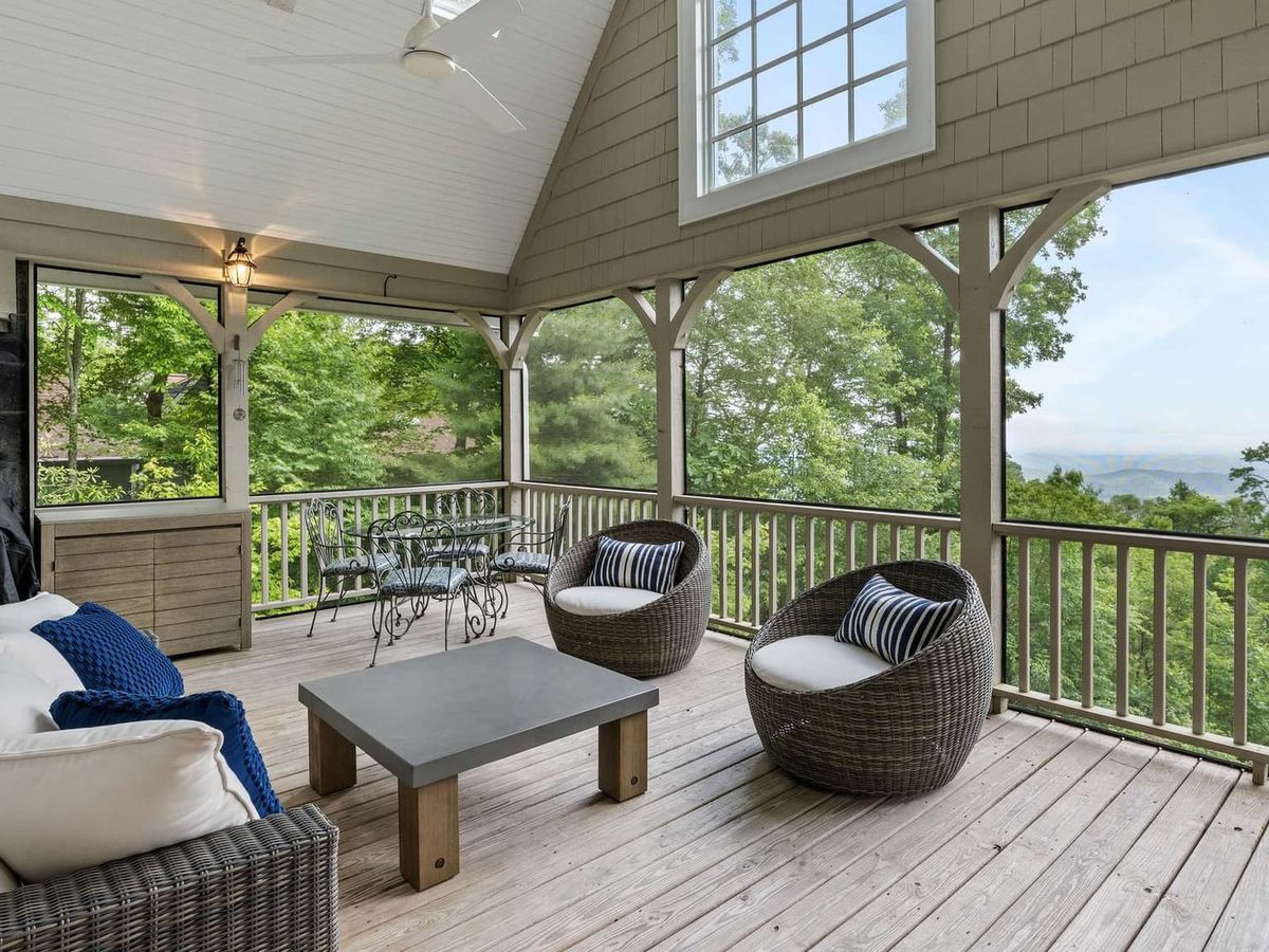 A covered mountain porch features cozy seating and a dining table with wide views of the forest and distant hills. This outdoor living space is ideal for morning coffee or quiet evenings surrounded by nature.
