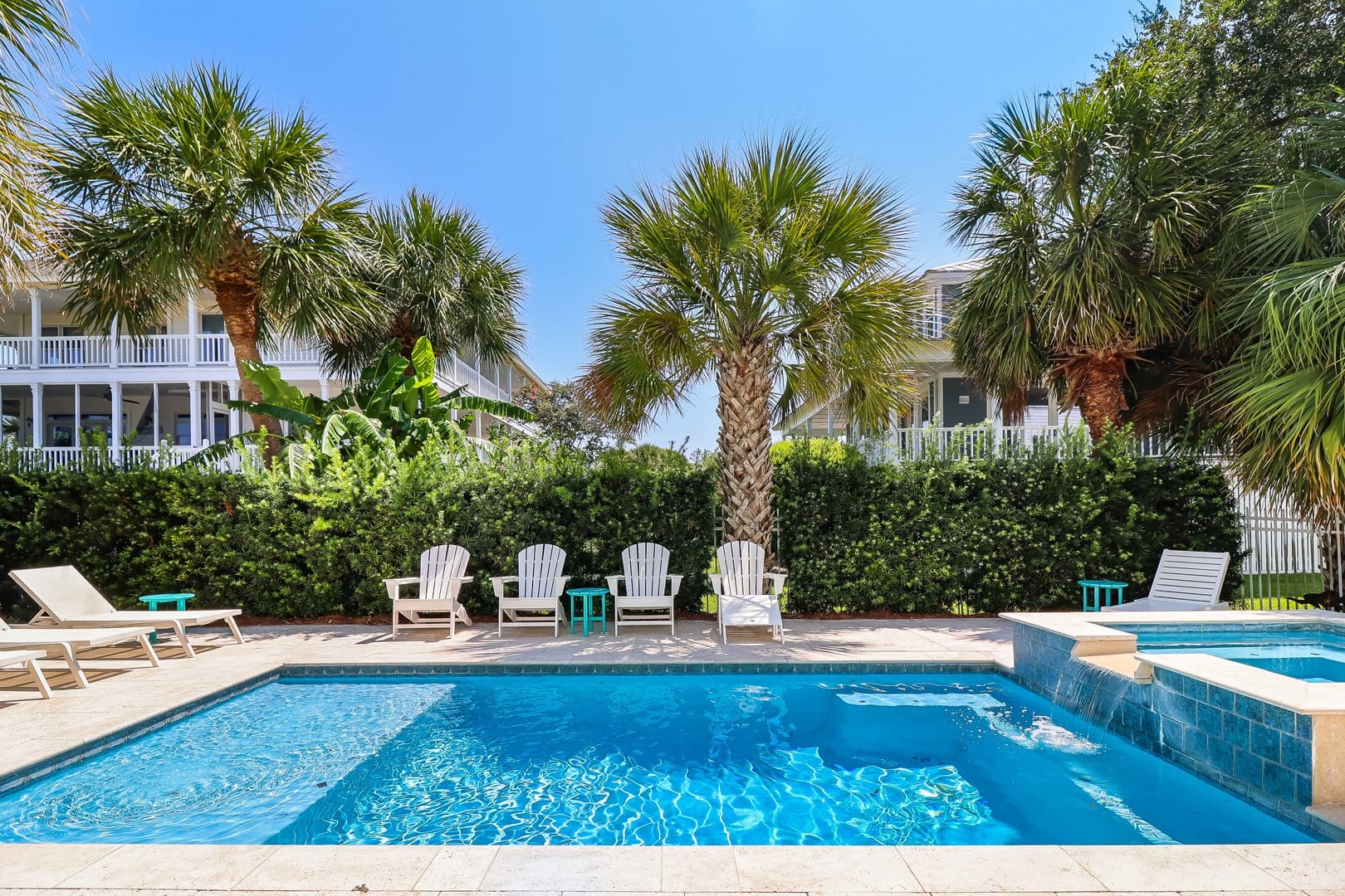 A resort-style pool and spillover hot tub at 7 Palmwood Court sits surrounded by white Adirondack chairs, lush palm trees, and trimmed hedges, with a coastal home's wraparound porches visible in the background. The sparkling blue water and tropical landscaping create an ideal private oasis for a beach vacation rental.