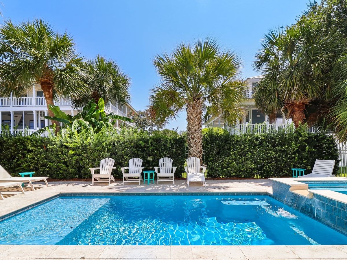 A resort-style pool and spillover hot tub at 7 Palmwood Court sits surrounded by white Adirondack chairs, lush palm trees, and trimmed hedges, with a coastal home's wraparound porches visible in the background. The sparkling blue water and tropical landscaping create an ideal private oasis for a beach vacation rental.