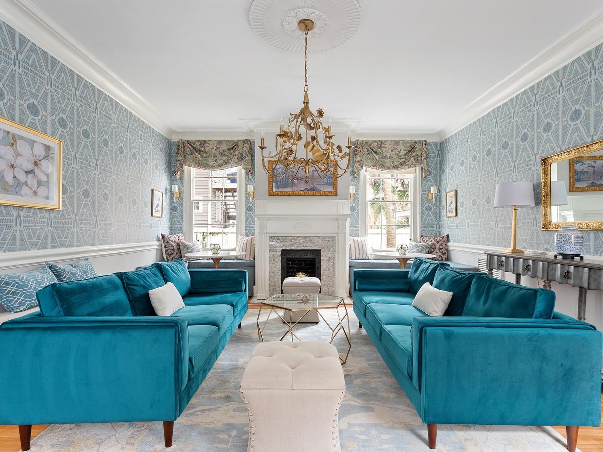 An elegant living room features two teal velvet sofas, a white fireplace, a gold chandelier, and patterned blue wallpaper with gold accents, creating a luxurious and classic atmosphere.