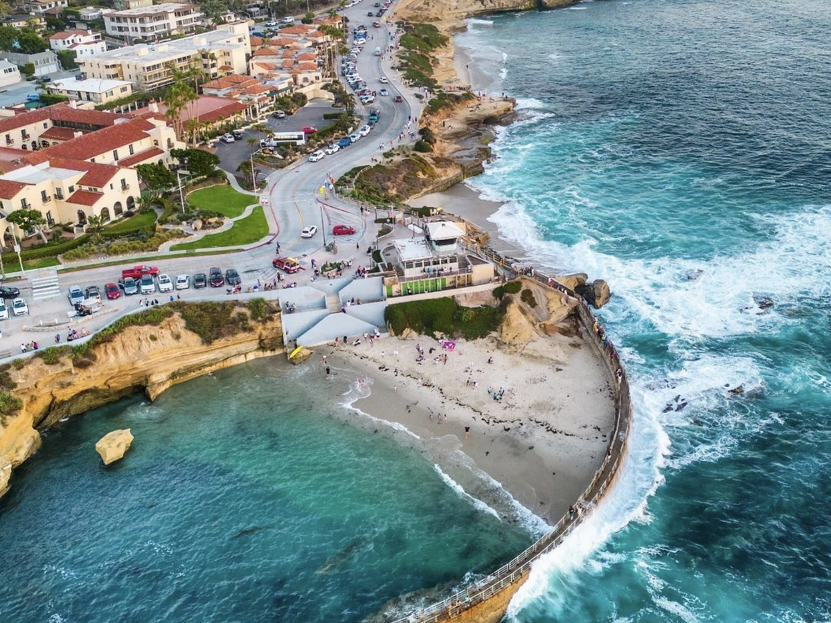 An aerial view of a coastal town with a curved seawall enclosing a sandy beach, turquoise waves crashing against the rocks, and people gathered along the shore and boardwalk.