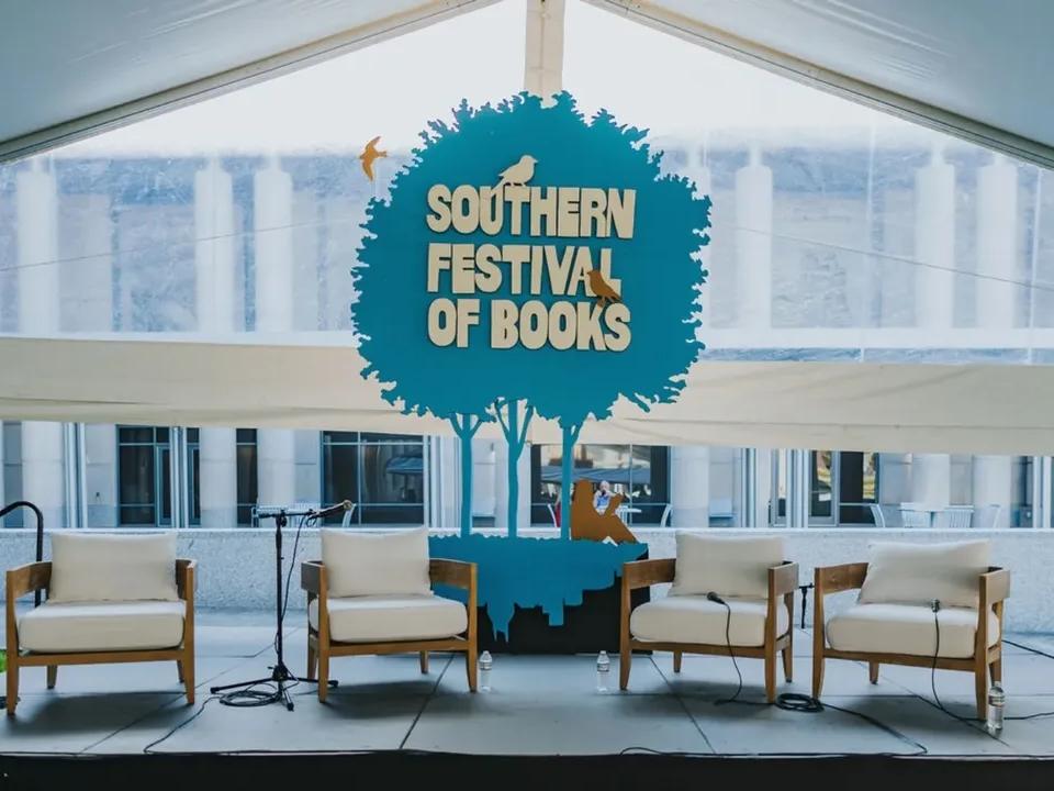 A clean, modern stage setup with cozy white chairs sits under a tent at the Southern Festival of Books. The large blue tree-shaped sign creates a fun and welcoming backdrop for author talks. It feels like the perfect spot to hear great stories and meet your favorite writers.