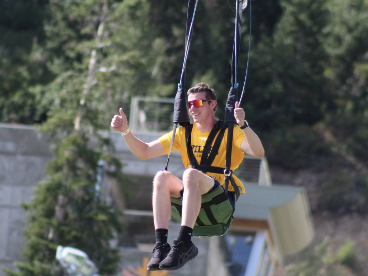 Man in a yellow shirt and sunglasses gives two thumbs up while riding a zipline chair through a forested mountain area.