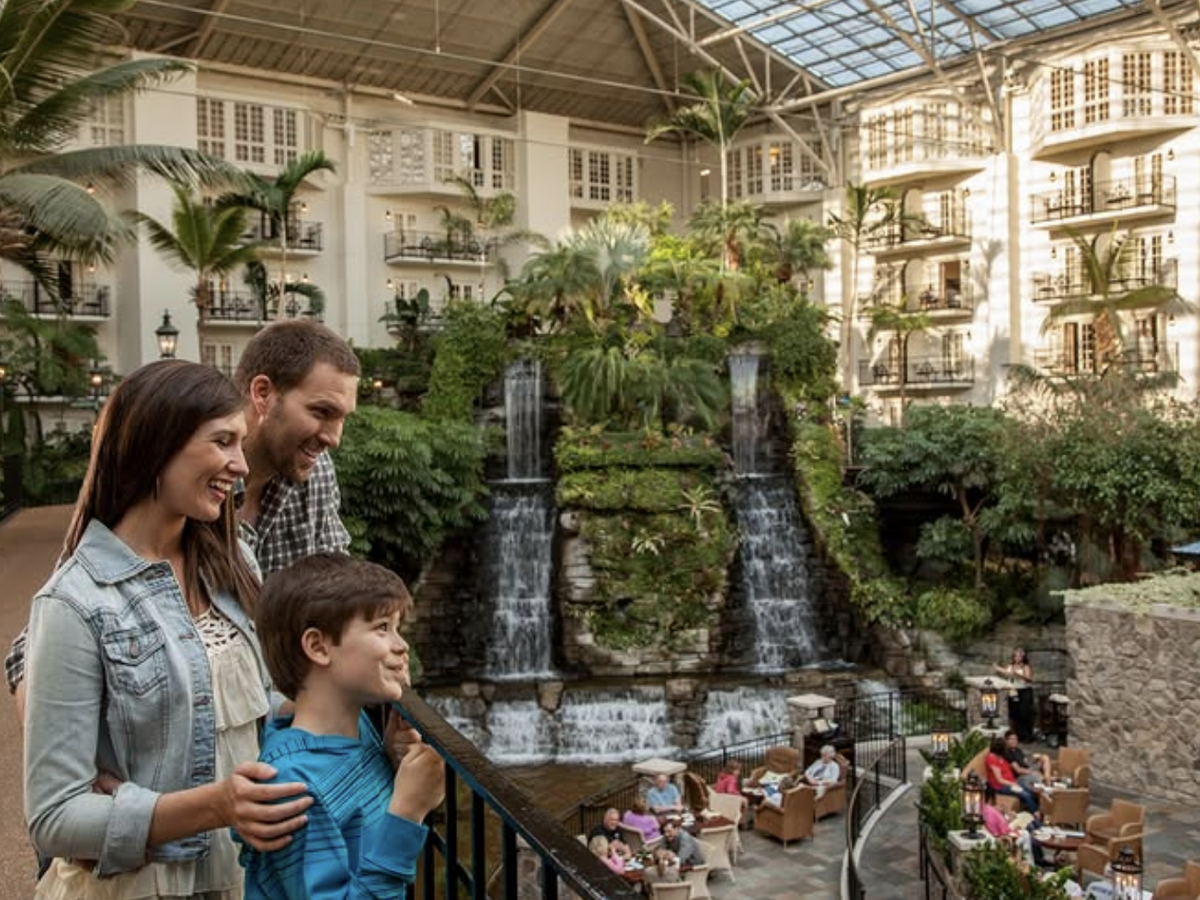 A smiling family enjoys the view of indoor waterfalls and lush greenery inside a grand hotel atrium with balconies and seating areas.
