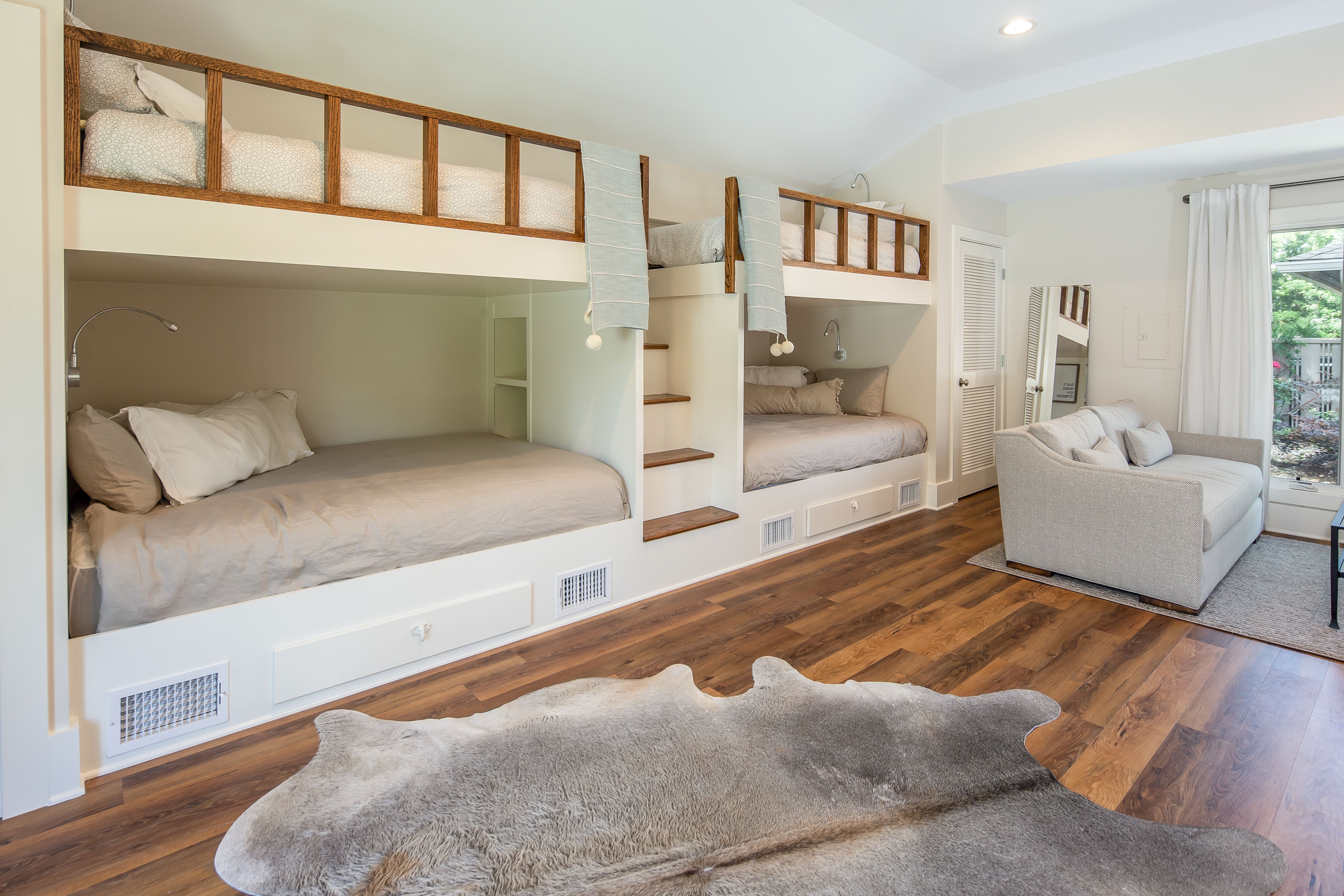 A stylish bunk room features two built-in lower beds and two upper loft beds with wooden railings and a central staircase, complemented by a cozy loveseat, soft lighting, and warm wood flooring with a faux fur rug.