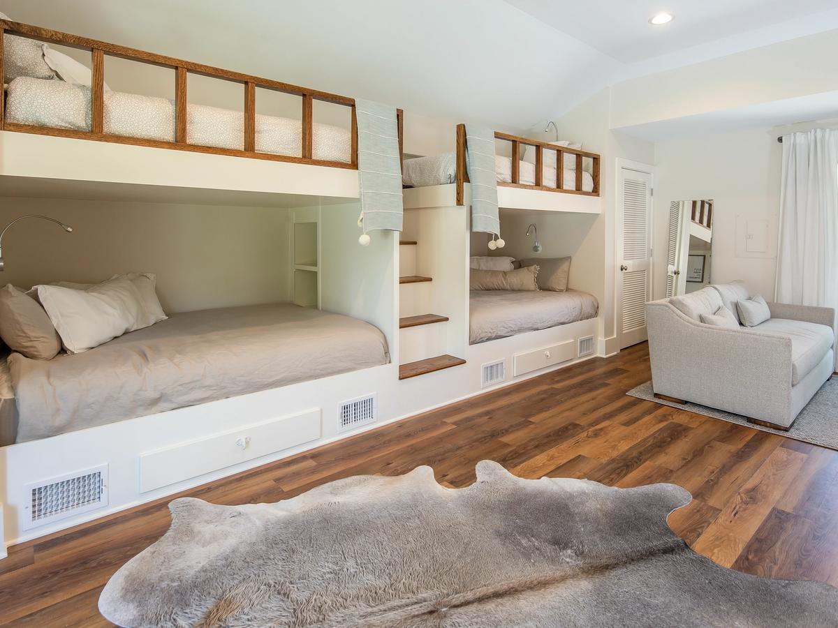A stylish bunk room features two built-in lower beds and two upper loft beds with wooden railings and a central staircase, complemented by a cozy loveseat, soft lighting, and warm wood flooring with a faux fur rug.