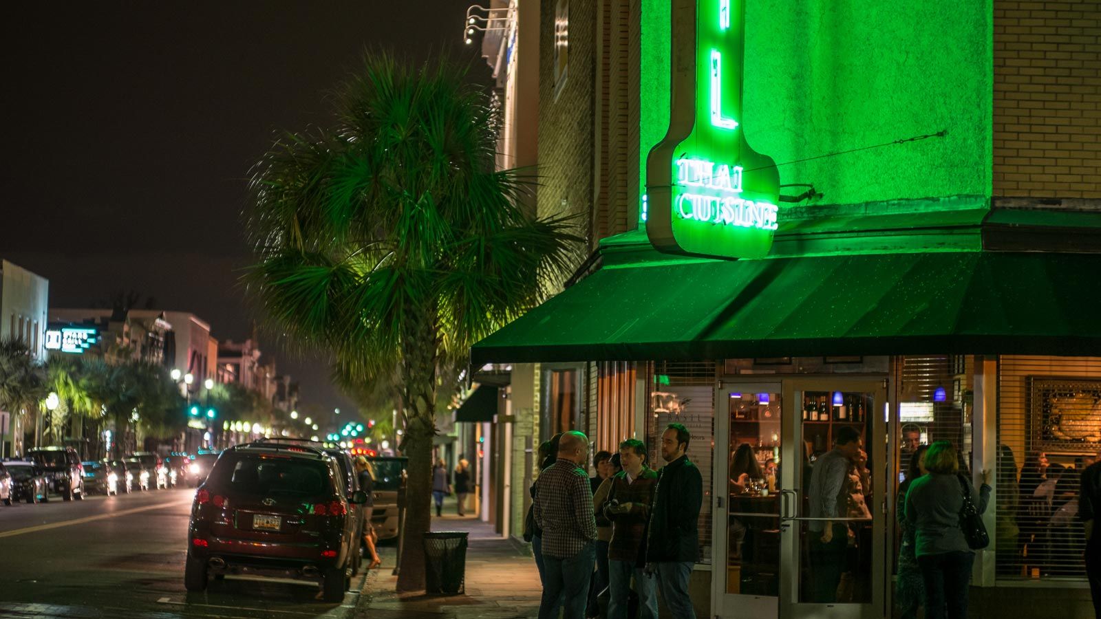A lively Charleston street at night glows with neon lights as people gather outside a Thai restaurant. Palm trees line the sidewalk while cars fill the road, giving the scene an energetic city vibe.