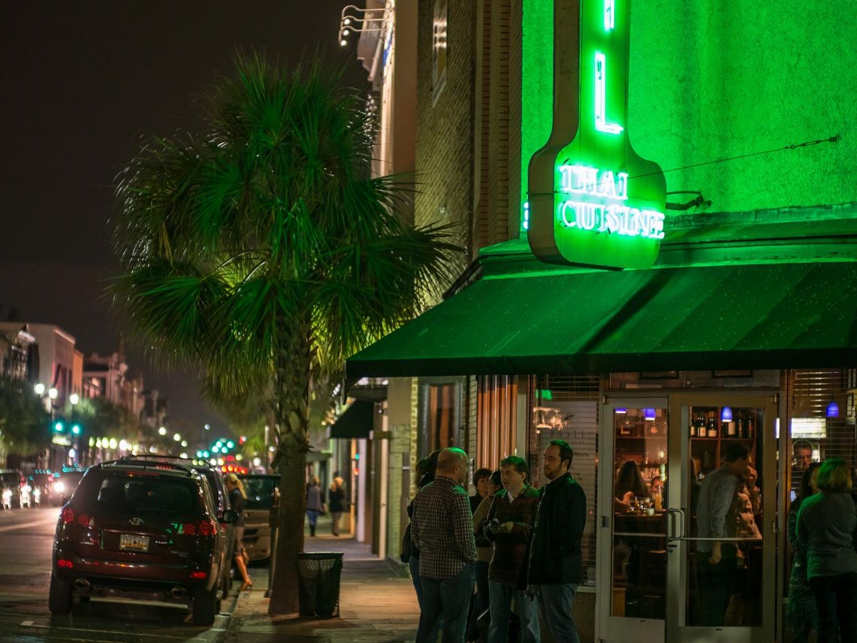 A lively Charleston street at night glows with neon lights as people gather outside a Thai restaurant. Palm trees line the sidewalk while cars fill the road, giving the scene an energetic city vibe.