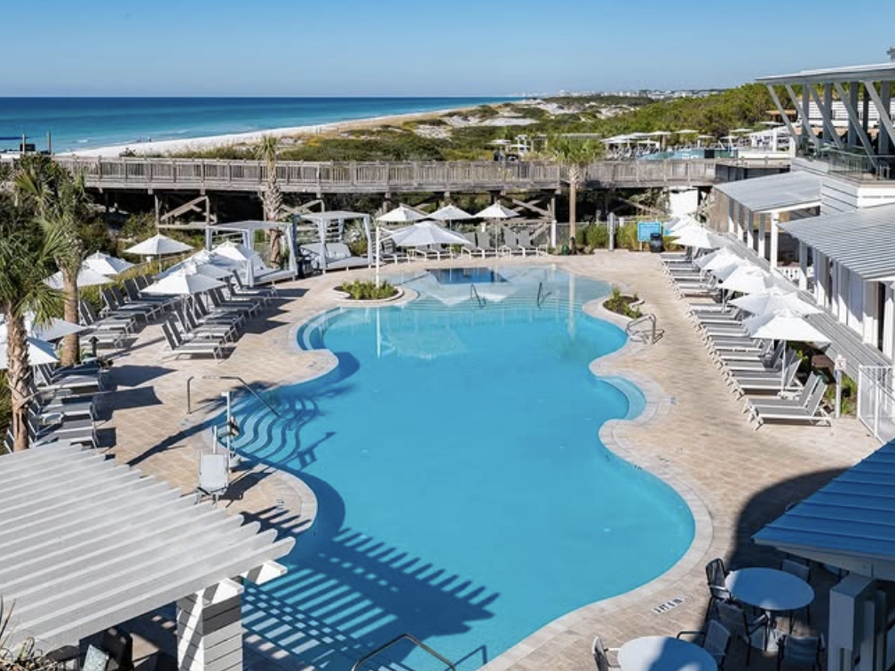 A resort-style pool surrounded by lounge chairs and white umbrellas, with palm trees and a wooden boardwalk leading to the white-sand beach and turquoise ocean in the background.