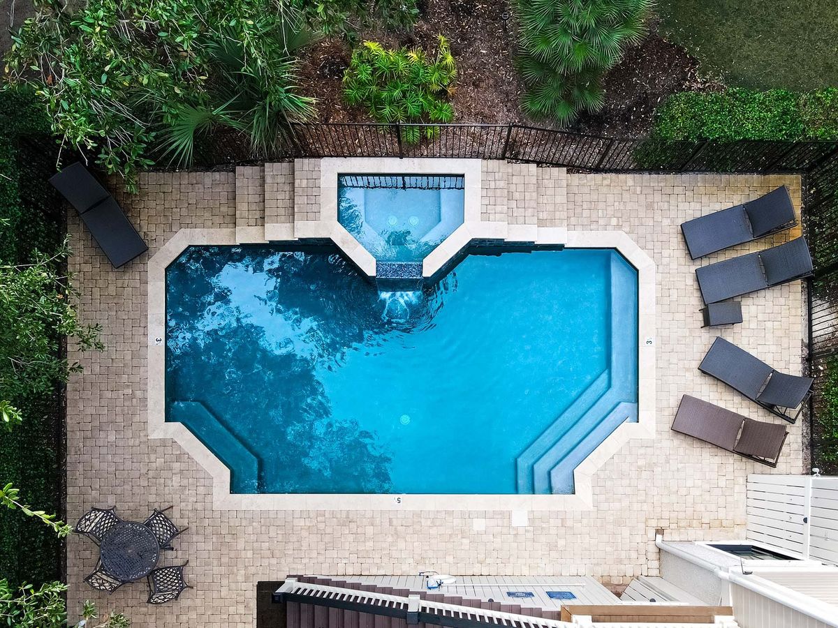 A luxurious outdoor pool area with a geometric design, featuring an attached elevated spa. The space is surrounded by lush greenery and a paved patio with lounge chairs and a bistro table, creating a private and relaxing retreat