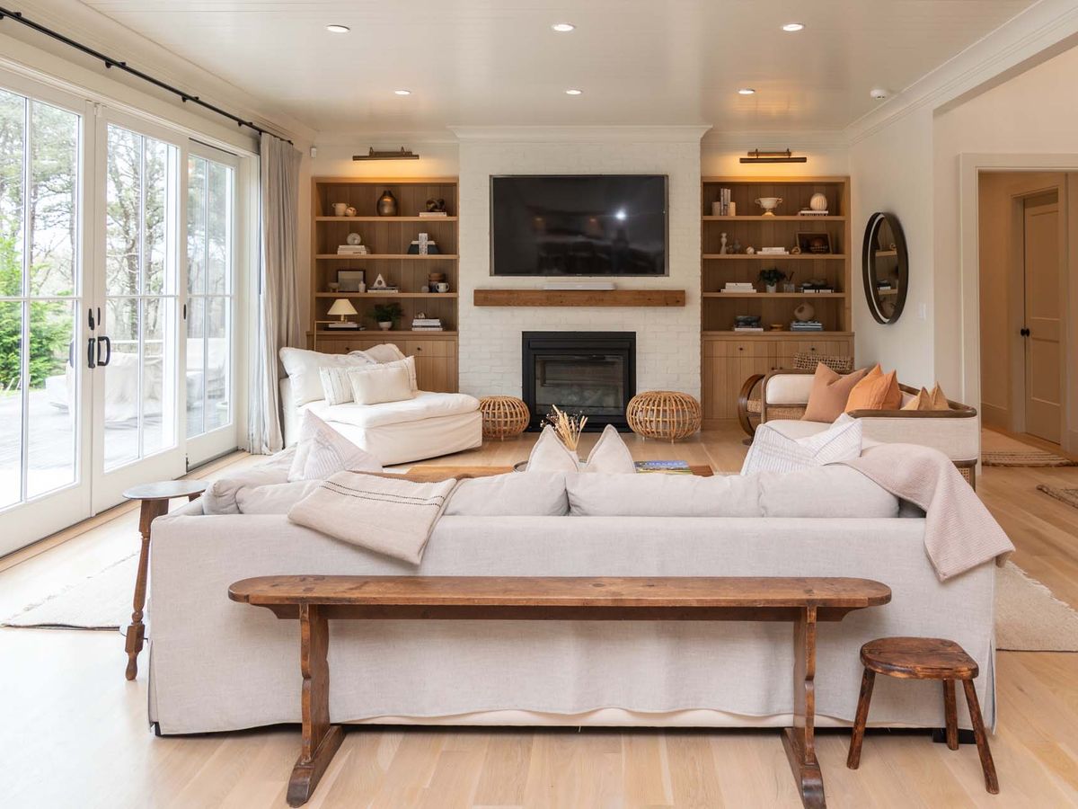 A cozy, neutral-toned living room features a white brick fireplace with a mounted TV, built-in wooden shelves, soft seating, and large sliding glass doors that open to an outdoor patio.