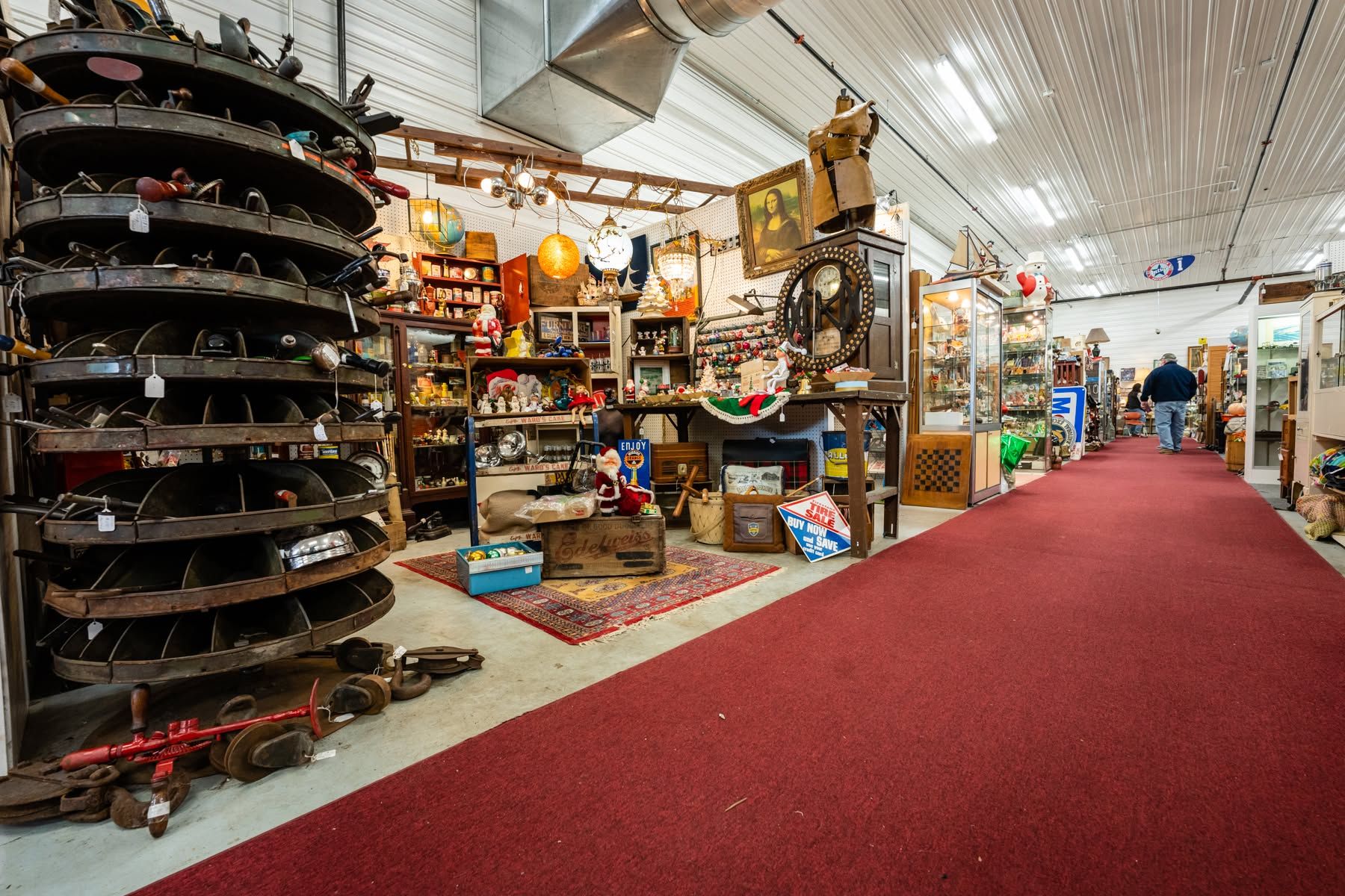 The interior of the Saugatuck Antique Pavilion stretches down a long red-carpeted aisle lined with crowded vendor booths full of vintage treasures, from a framed Mona Lisa print to old signs, holiday decor, and glass display cases. A shopper browses the eclectic collection under bright industrial lighting.