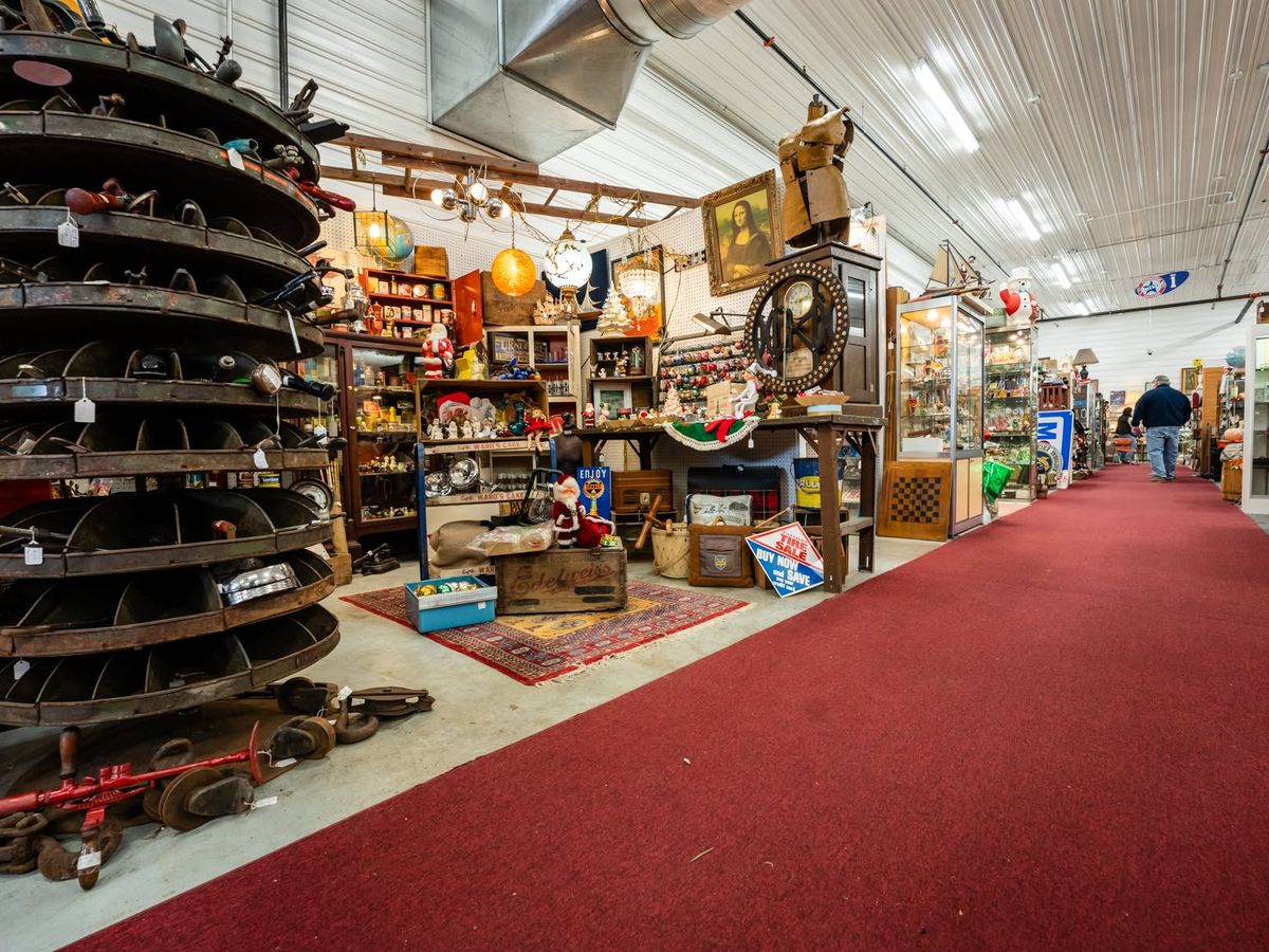 The interior of the Saugatuck Antique Pavilion stretches down a long red-carpeted aisle lined with crowded vendor booths full of vintage treasures, from a framed Mona Lisa print to old signs, holiday decor, and glass display cases. A shopper browses the eclectic collection under bright industrial lighting.