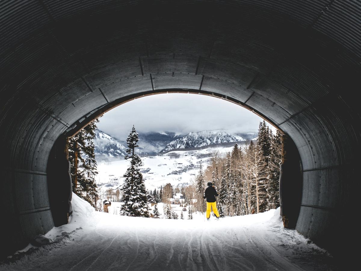 A skier in bright yellow pants stands at the edge of a snowy tunnel, gazing out toward a dramatic mountain valley blanketed in fresh powder and framed by snow-covered trees.