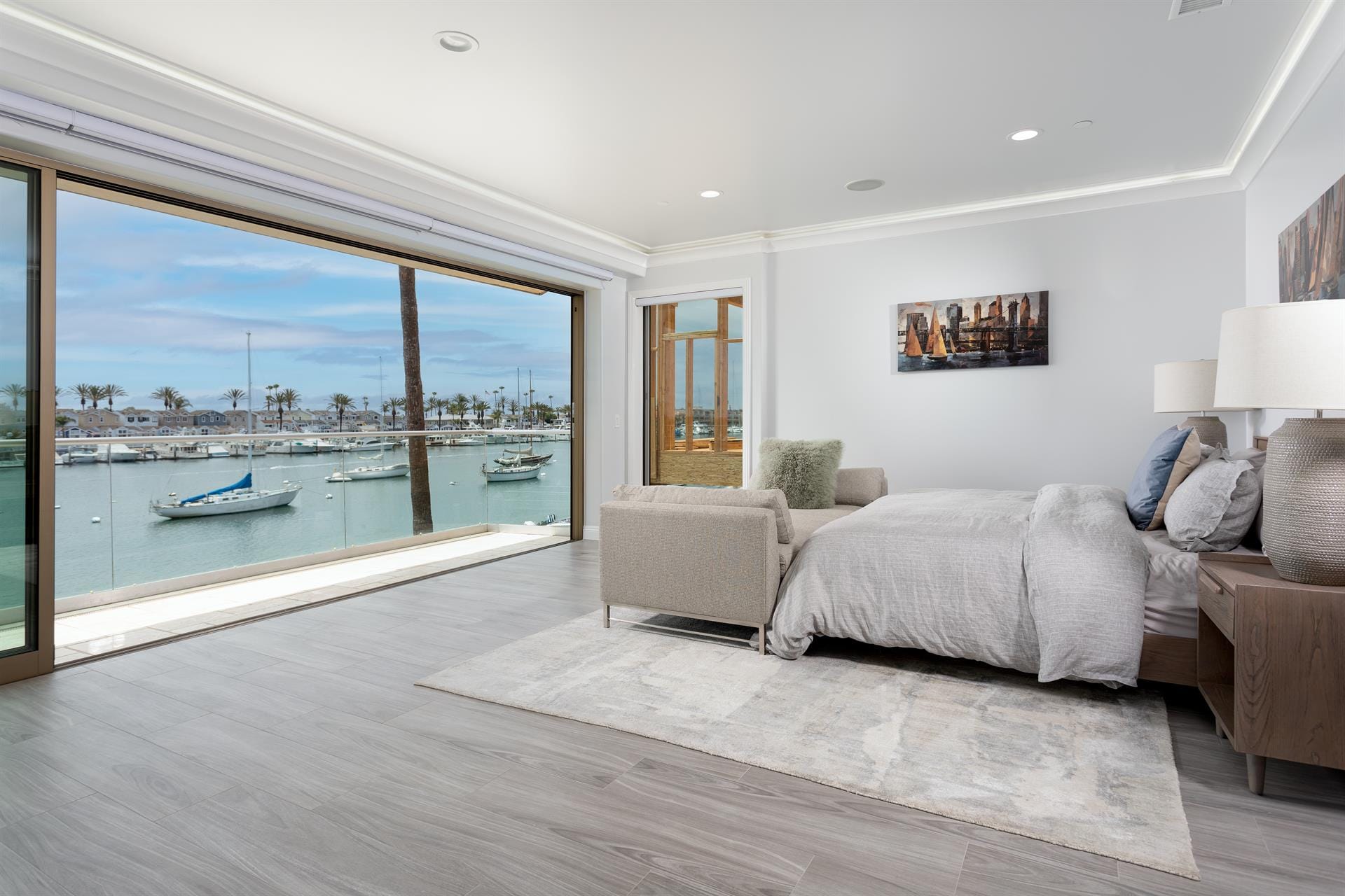 A modern waterfront bedroom features a wide glass wall with stunning views of boats and the marina. Soft neutral tones and clean design create a relaxing coastal escape.