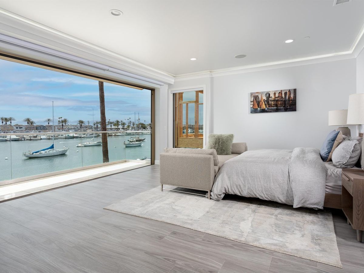 A modern waterfront bedroom features a wide glass wall with stunning views of boats and the marina. Soft neutral tones and clean design create a relaxing coastal escape.