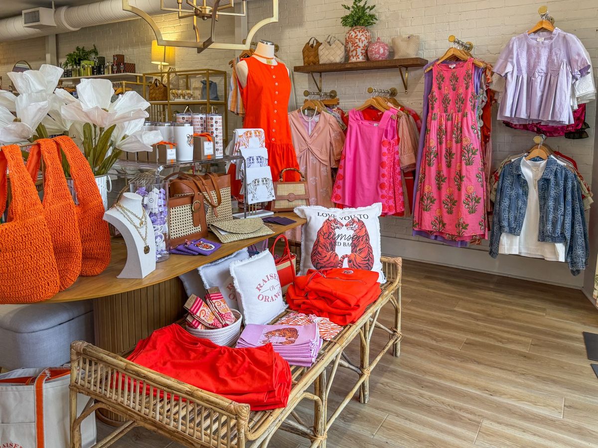 True Orange Boutique showcases a vibrant mix of orange and pink Clemson-spirited apparel, including dresses, graphic tees, and tiger-motif throw pillows arranged on a rattan bench and display table. White brick walls, warm wood shelving, and gold fixtures give the cheerful shop a polished, welcoming feel. 