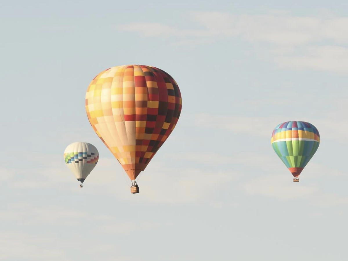 Three colorful hot air balloons float peacefully in a pale blue sky scattered with soft clouds.