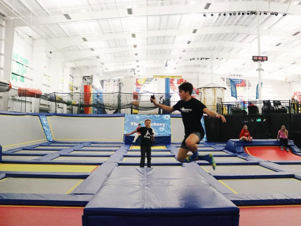 Kids jump and play on interconnected trampolines inside a large, brightly lit indoor trampoline park.