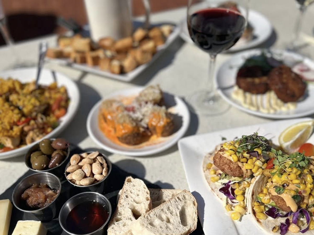 Outdoor table spread with red wine, fish tacos topped with corn salsa, meatballs in red sauce, charcuterie board with olives and nuts, and assorted tapas-style small plates.