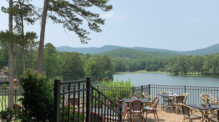 A cozy outdoor dining area at Big Canoe, GA, overlooking a peaceful lake framed by lush pine trees and rolling mountains. The patio’s wicker chairs and tables create a relaxed atmosphere, perfect for enjoying meals with a view of the Blue Ridge landscape