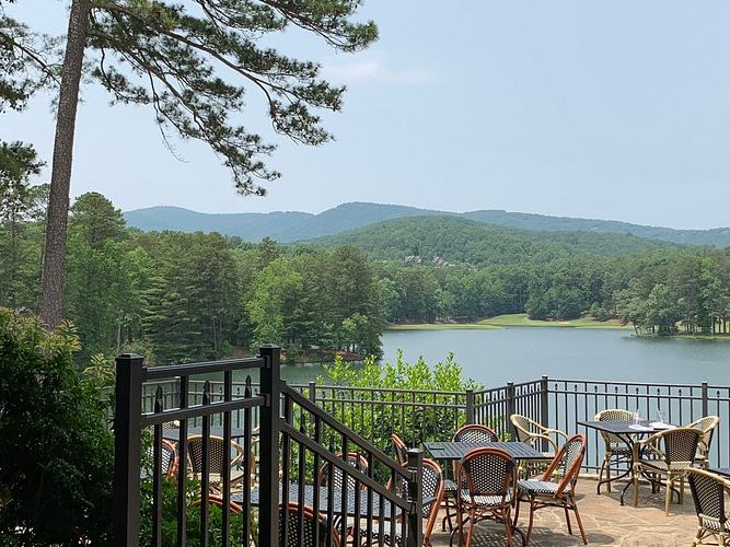 A cozy outdoor dining area at Big Canoe, GA, overlooking a peaceful lake framed by lush pine trees and rolling mountains. The patio’s wicker chairs and tables create a relaxed atmosphere, perfect for enjoying meals with a view of the Blue Ridge landscape