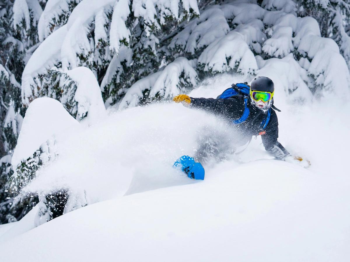 Man in blue snow gear and helmet snowboarding on deep powedered snow with trees behind him
